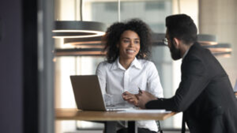 A professional woman and man sitting at a desk, collaborating and exchanging looks, with a laptop in front of them