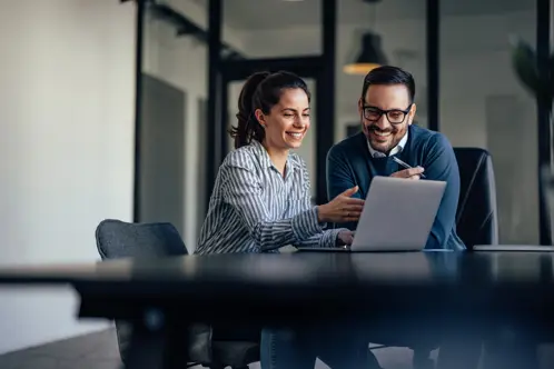 Una donna e un uomo, seduti a una scrivania, sorridenti e guardando lo schermo di un laptop