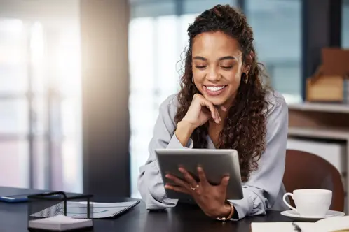 Una donna seduta alla sua scrivania, sorridente mentre guarda lo schermo di un iPad, con una mano appoggiata sul mento e l'altra che tiene il dispositivo