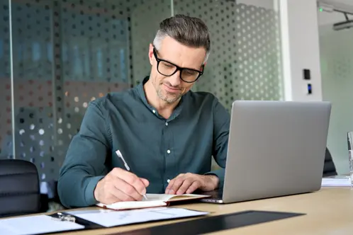 Un homme avec des lunettes assis  un bureau, souriant en crivant dans un carnet