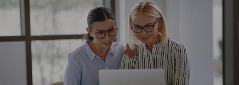 Two professional women with glasses, smiling as they look at a laptop screen