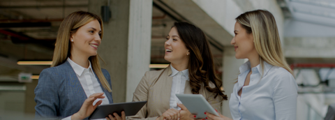Three professional women standing together, each holding an iPad and smiling warmly at one another