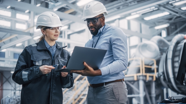 A man and a woman in hard hats and safety glasses holding a laptop, ready to work on a construction site