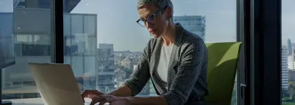 Focused businesswoman concentrating on her laptop in a modern office with large windows overlooking a cityscape, working.