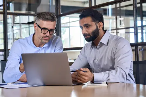 Two professional men seated side by side, focused on a laptop screen as they collaborate intently