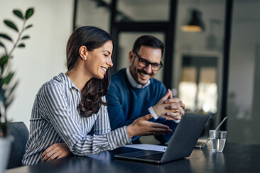 Two colleagues sitting at a table in a modern office, smiling and discussing something on a laptop screen, with a glass of water and a plant in the background.