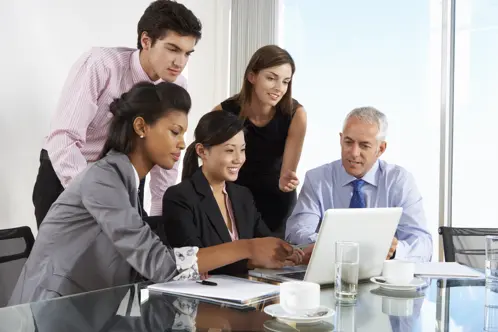 Un gruppo di professionisti in piedi insieme in una sala conferenze, riuniti intorno e guardando intensamente lo schermo di un laptop
