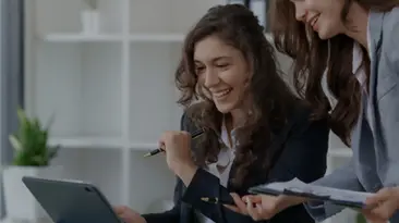 Eine Frau steht, whrend die andere sitzt, beide lcheln, whrend sie auf ihren Laptop-Bildschirm schauen
