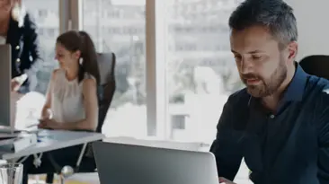 Ein berufsttiger Mann sitzt in einer Broumgebung und tippt mit ernster Miene auf seinem Laptop