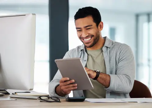 A man seated, holding an iPad and smiling as he looks at the screen