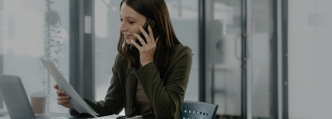 Een professionele vrouw die aan haar bureau in een kantoor zit, met een stuk papier in de ene hand en een mobiele telefoon aan haar oor met de andere, glimlachend terwijl ze praat