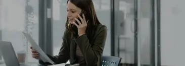 Una mujer profesional sentada en su escritorio en una oficina, sosteniendo un pedazo de papel en una mano y un telfono celular en su oreja con la otra, sonriendo mientras habla