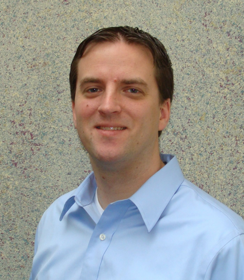 A professional man in a blue shirt and tie stands confidently in front of a plain wall.