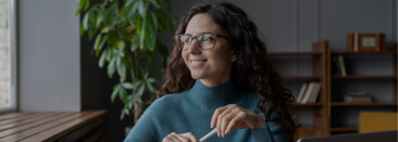 A woman wearing glasses holds a pen and smiles while gazing out the window