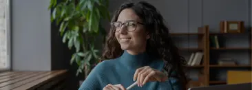 A woman wearing glasses holds a pen and smiles while gazing out the window