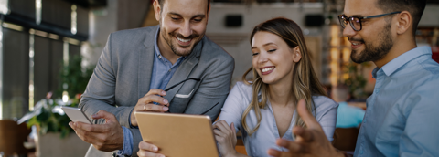 Three professionals stand together, smiling as they look at a laptop