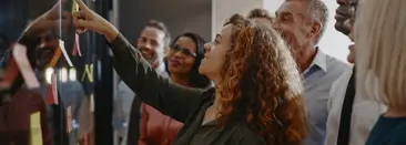 Een vrouw glimlacht terwijl ze wijst naar een bord bedekt met plakbriefjes, omringd door een groep lachende professionals
