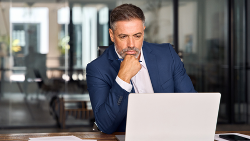 A man sits at his desk with a serious expression, resting one hand on his chin as he looks at his laptop screen