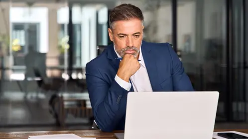 A man sits at his desk with a serious expression, resting one hand on his chin as he looks at his laptop screen