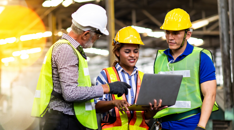 Three workers in hard hats and safety vests collaborating on a laptop at a construction site.