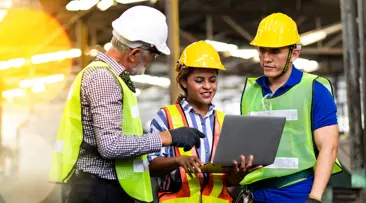 Three workers in hard hats and safety vests collaborating on a laptop at a construction site.
