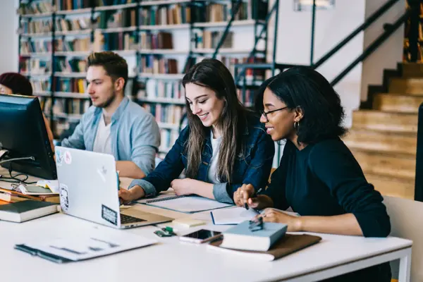 Three individuals collaborating on a laptop in a library, engrossed in their work