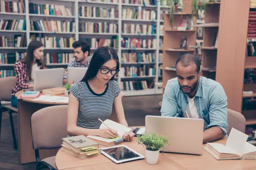 A diverse group of individuals sitting around a table in a quiet library, engrossed in their studies and surrounded by books