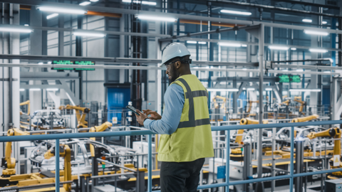 A man in a hard hat and vest stands in an industrial area, overseeing construction activities.