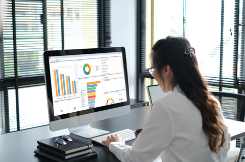 A woman in a white shirt sits at a desk, analyzing graphs on a computer screen.