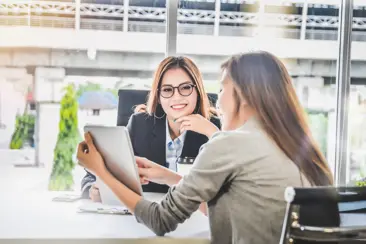 Twee professionele vrouwen zitten en glimlachen, terwijl de een de ander iets op het scherm van een iPad laat zien