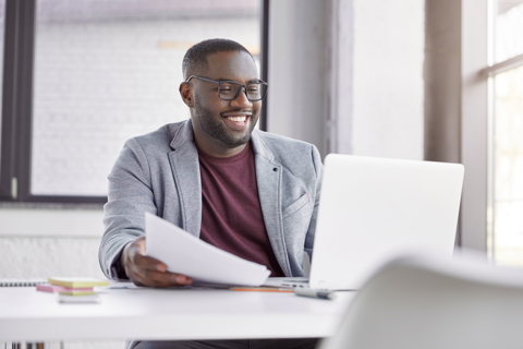 A professional black man with a cheerful expression sits at a desk, working on a laptop computer.