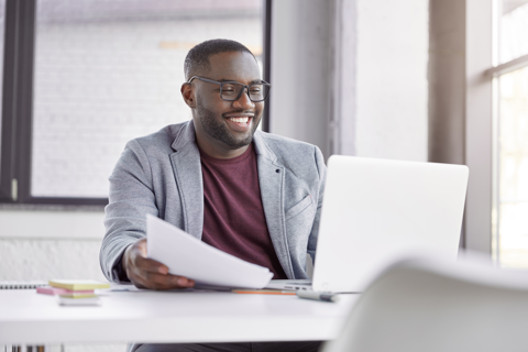 A professional black man with a cheerful expression sits at a desk, working on a laptop computer.