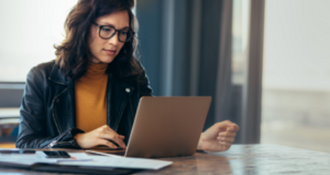 A woman wearing glasses sits at a table, focused on her laptop.