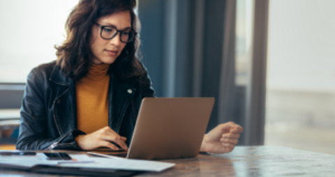 A woman wearing glasses sits at a table, focused on her laptop.