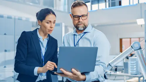 A man and woman in business attire focused on a laptop screen, engaged in professional work