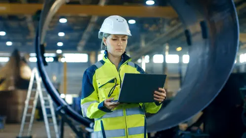 A woman in a hard hat and safety vest holding a laptop, ready to work on a construction site