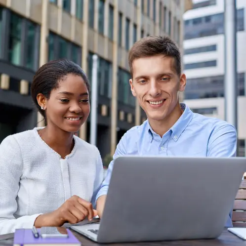 Deux professionnels assis ensemble l?extrieur d?un btiment, souriant devant leur ordinateur portable