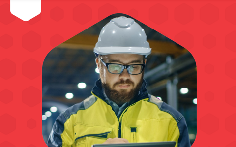 A man in a hard hat and safety glasses holding a tablet, wearing a yellow jacket, ready to work on a construction site