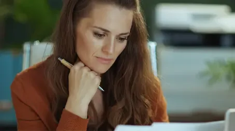 A woman sits with a pen in hand, smiling as she looks at a paper, with a laptop in front of her