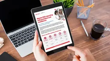 Eine Person hlt ein iPad mit Text in der Hand, vor sich einen Laptop und daneben eine Kaffeetasse
