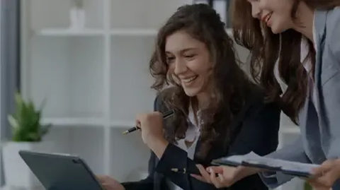 Uma mulher fica de p enquanto a outra se senta, ambas sorrindo enquanto olham para a tela do laptop
