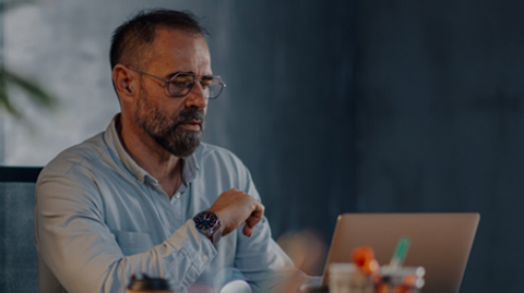 A professional man with glasses sits and looks at his laptop screen with a serious expression