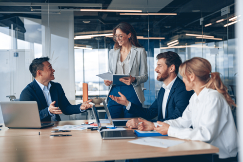 Group of business professionals in a modern office having a collaborative meeting, smiling and discussing ideas, with laptops and documents on the table.