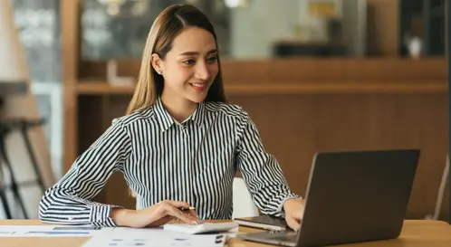 Une femme en blouse raye, assise  son bureau, souriante en regardant l?cran de son ordinateur portable