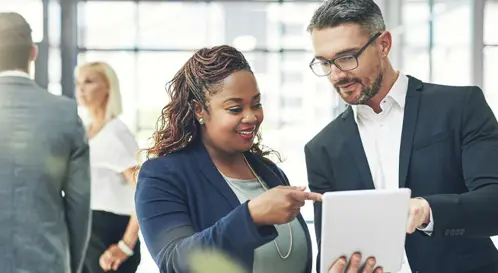 Une femme et un homme professionnels debout dans un bureau, souriant et collaborant en pointant un morceau de papier qu?ils tiennent ensemble