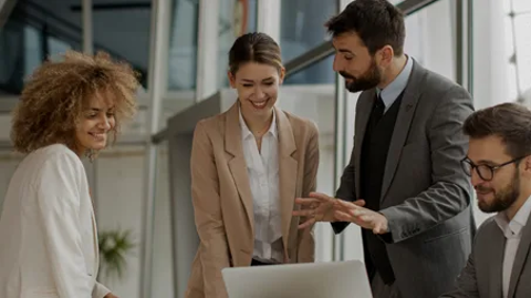 A group of business people gathered around a laptop, smiling, and engaged in a discussion or collaboration