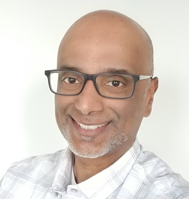 A close-up of a man with glasses, smiling and showing his teeth
