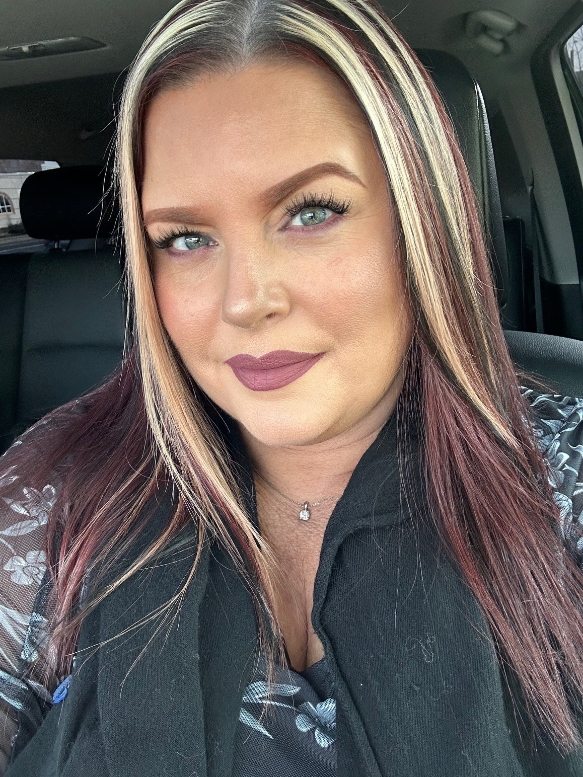 A woman with blonde and purple hair, sitting in her car and smiling