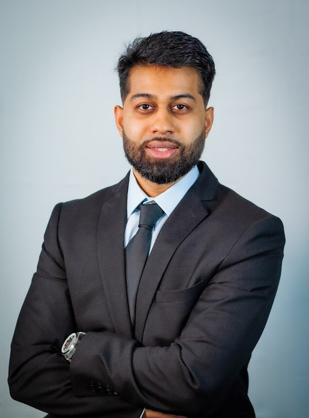A man in a suit, standing with his arms crossed and a subtle smile on his face