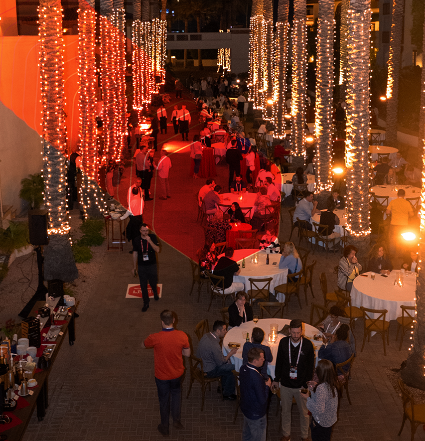A large networking event featuring tables and a buffet, with numerous professionals standing and sitting, engaged in conversation under ambient lighting